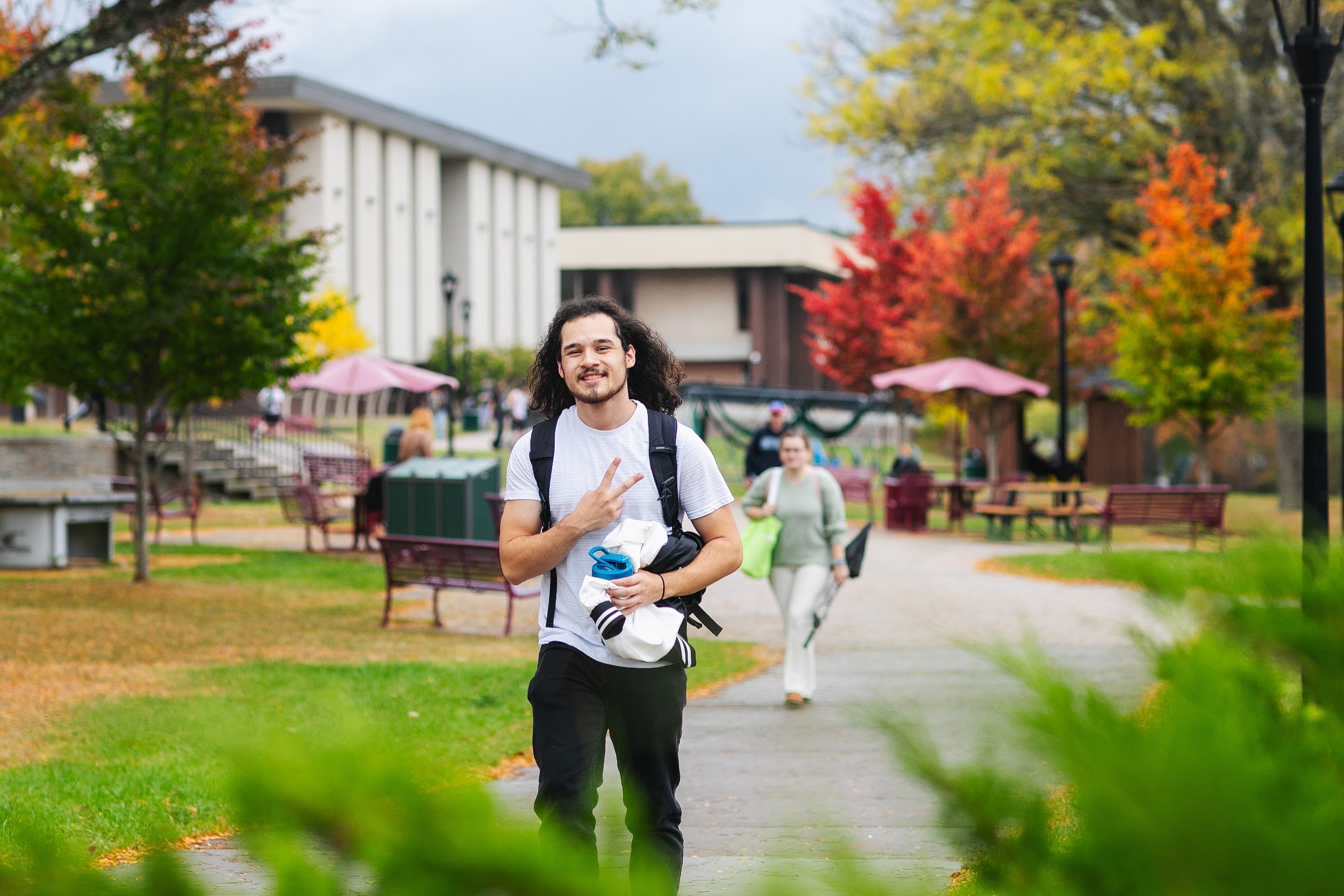 A smiling student walks on a leafy campus path, holding clothes and flashing a peace sign.